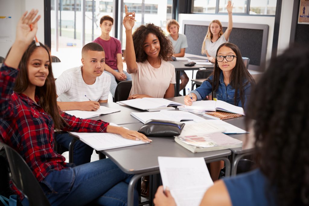 High,School,Kids,Raise,Hands,,Teacher,Sitting,At,Their,Desk